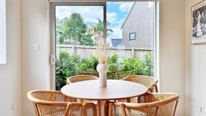 Ein Essbereich mit einem runden Tisch und sechs Korbstühlen, einer weißen Vase mit Trockenpflanzen sowie Blick auf einen Garten mit Holzzaun und Grünpflanzen.