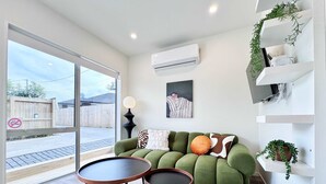 Ein modernes Wohnzimmer mit einem grünen Sofa, zwei runden Tischen, einem weißen Wandregal mit Pflanzen und einem großen Fenster mit Blick auf einen Holenzaun.