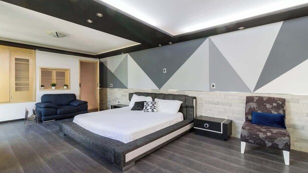 A modern bedroom with a large bed, a dark wood floor, a grey and white geometric wall design, a black nightstand, a blue sofa, and a patterned armchair. - Mini depa loft (Zapopan)