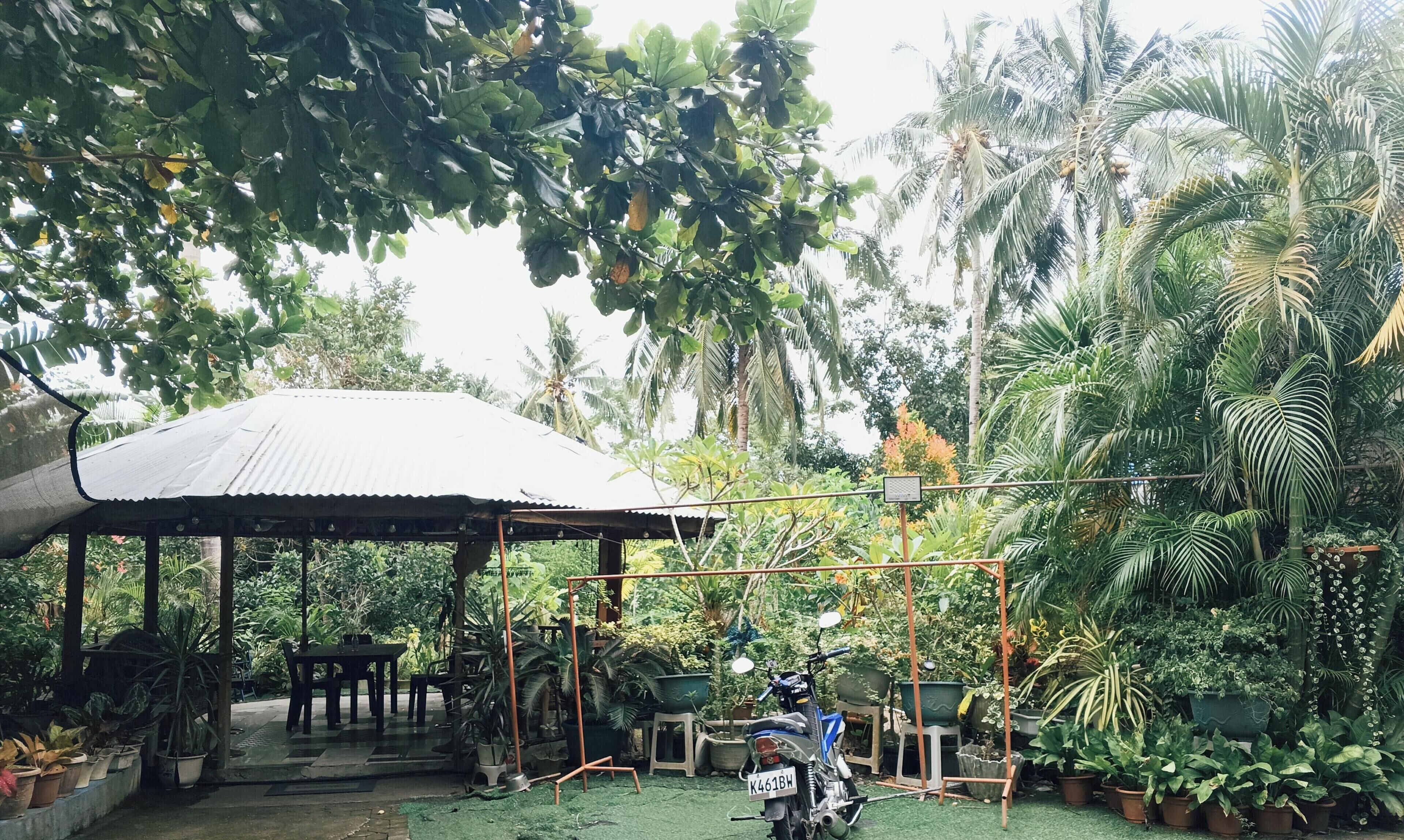 A covered outdoor seating area with a motorcycle parked nearby, surrounded by lush greenery and potted plants.