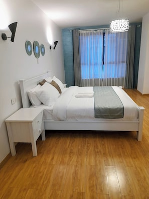 Een moderne slaapkamer met een groot bed, witte nachtkastjes, een houten vloer en een plafondlamp.