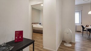 A modern hotel room with a bed, a small table with a red welcome sign, a white toilet, and a dining area with a table and chairs. - Quinto Modern Flat by Wonderful Italy (Genoa)