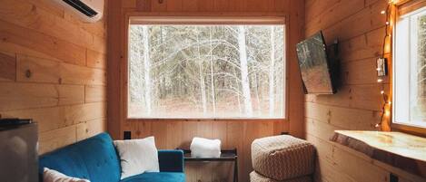 Interior acogedor de una cabaña con un sofá azul, una mesita, una ventana con vistas a árboles y un techo de madera.
