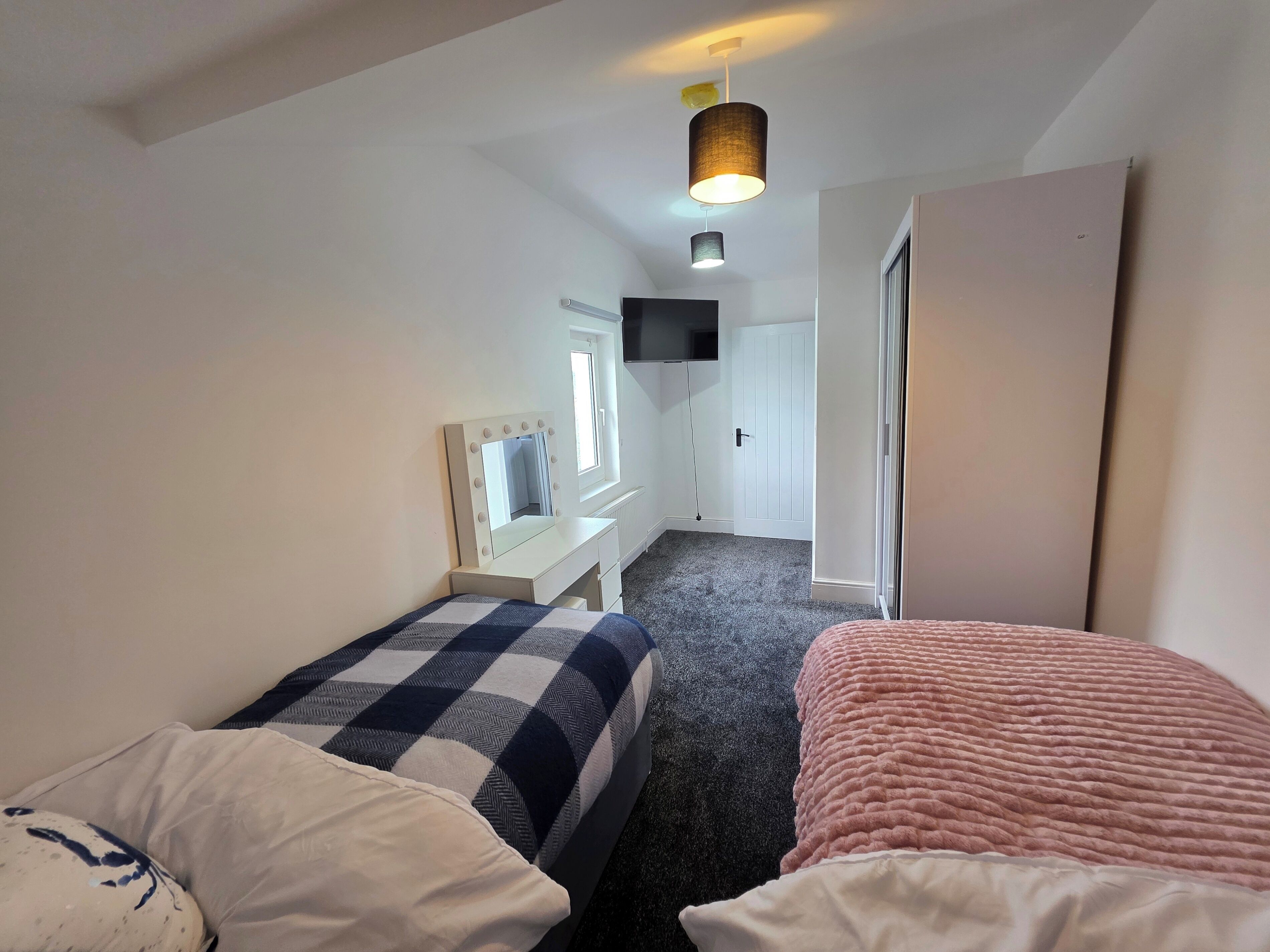 Une chambre avec deux lits, une coiffeuse avec un miroir, un téléviseur fixé au mur et une porte donnant sur une autre pièce.