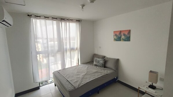 Een slaapkamer met een bed, een raam met gordijnen, een wandairconditioner en een klein tafeltje met een lamp.
