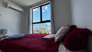 A bedroom with a large window, a bed with a red blanket, and a view of a cityscape and mountain.