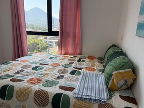 Un lit recouvert d’une courtepointe à motifs, avec des rideaux roses et une vue sur des bâtiments et des montagnes par la fenêtre.