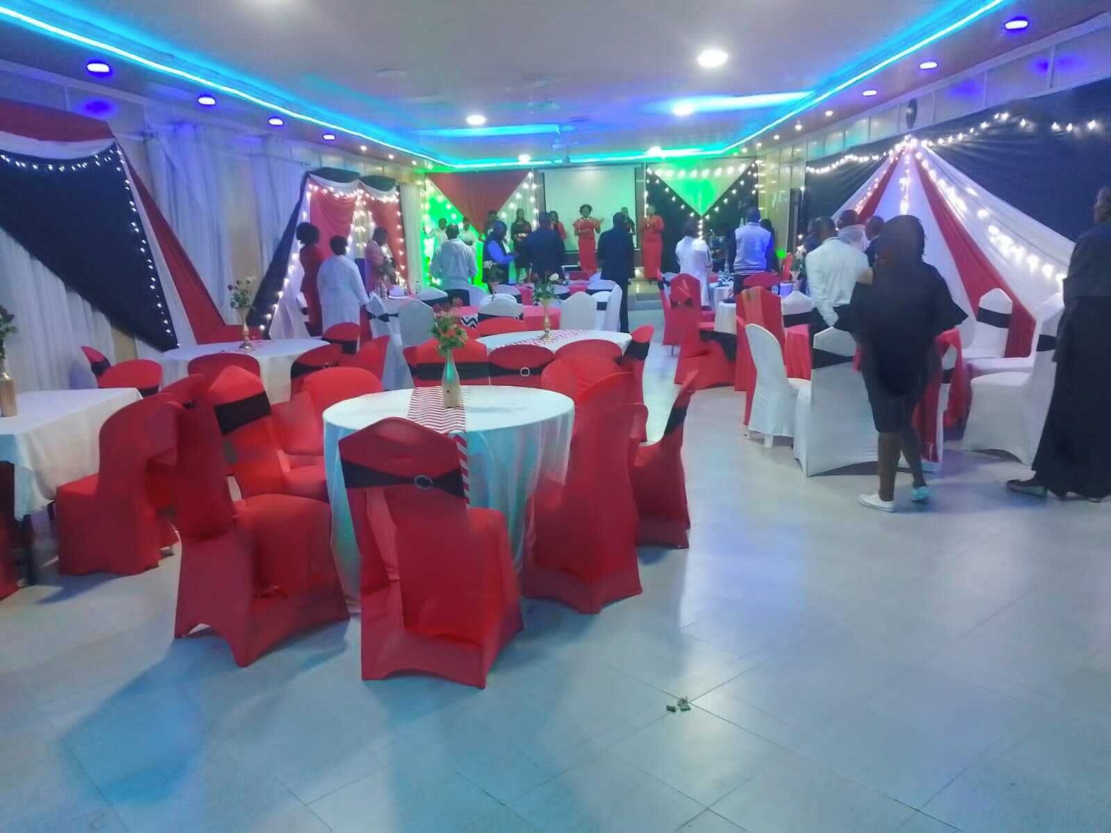 A banquet hall with round tables and red chairs, decorated with white and red drapes, and string lights.