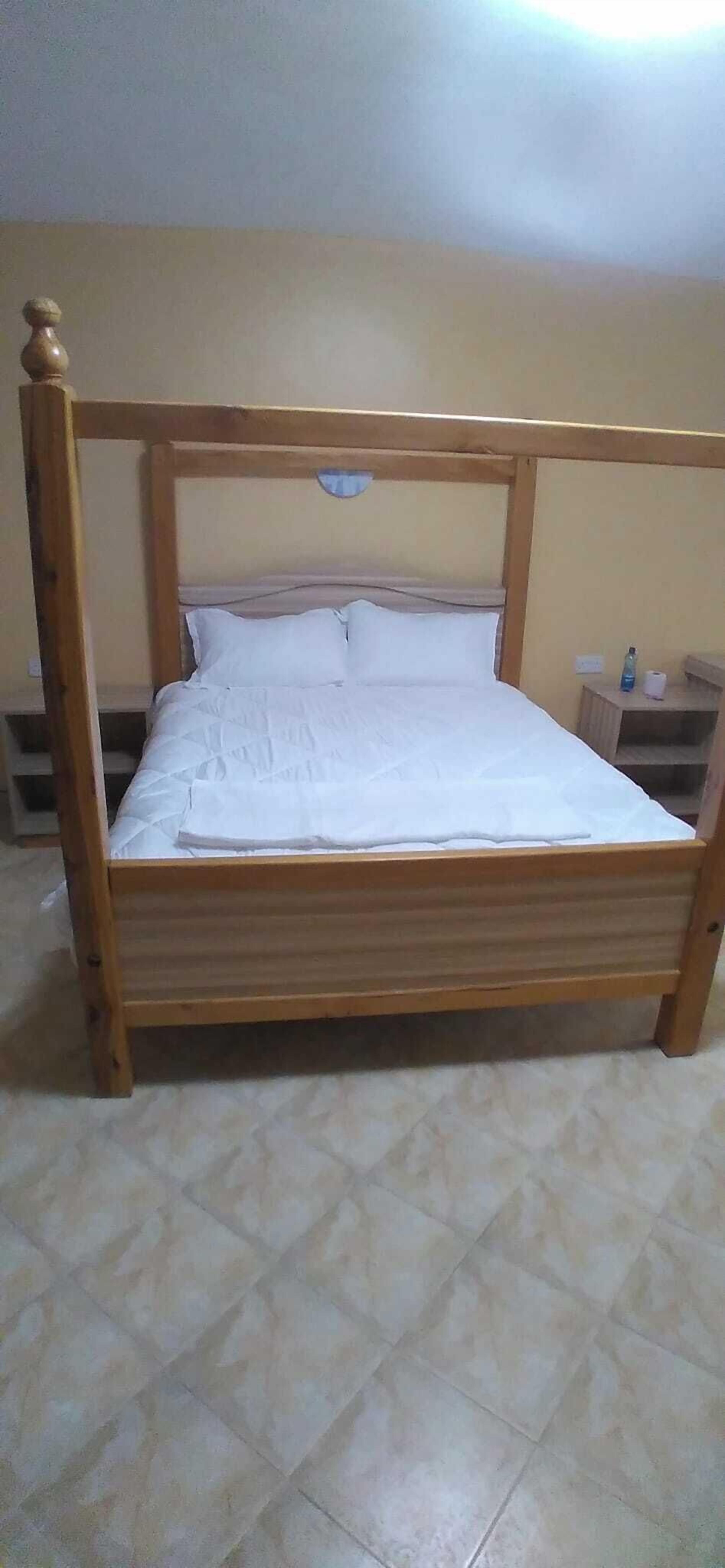 Una cama de madera con cabecera, ropa de cama blanca y una mesita de noche con una botella y una taza.