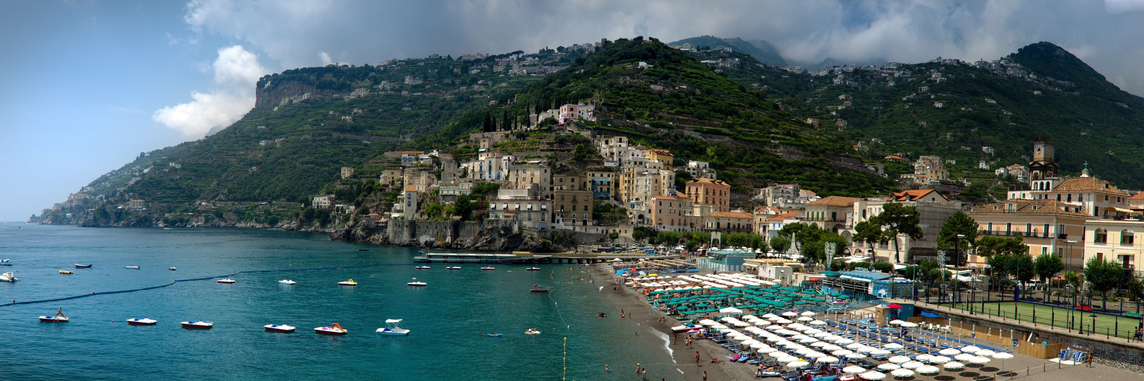 A coastal town with a beach, boats, and buildings on a hillside.