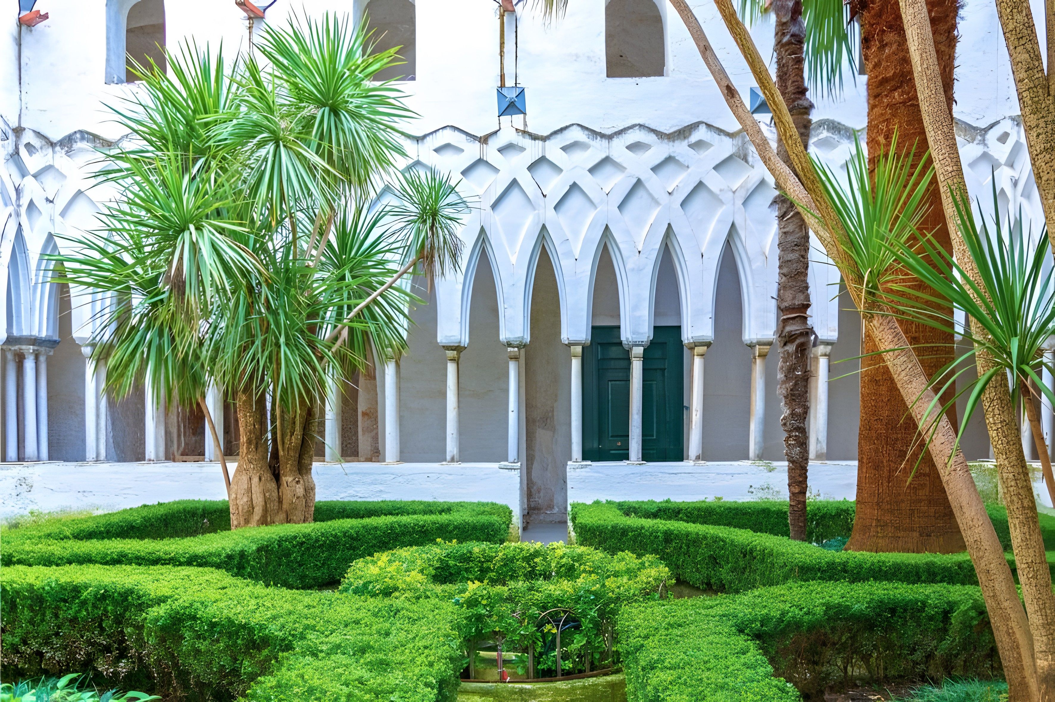 A courtyard with a white building featuring arched doorways and columns, surrounded by well-maintained greenery and palm trees.