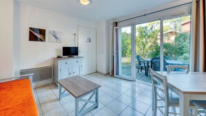 A modern, bright room with a dining table, chairs, a TV, and a sliding glass door leading to an outdoor area with a table and chairs.