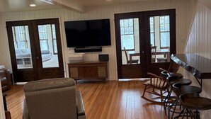 A spacious living room with a large flat-screen TV, wooden flooring, a sofa, and a dining area with chairs.