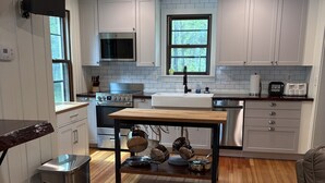 A modern kitchen with white cabinets, a wooden dining table, stainless steel appliances, and a window with a view of trees.