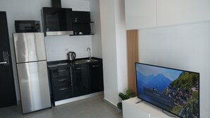 A modern kitchen with a stainless steel refrigerator, black cabinets, a kettle, and a sink. A flat-screen TV is mounted on the wall displaying a scenic image.