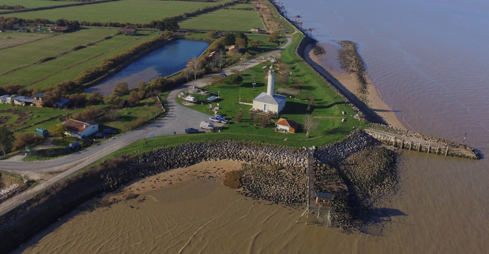 An aerial view of a coastal area with a lighthouse, a small harbor, and a stone breakwater.