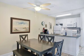 A dining area with a table and chairs, a ceiling fan, a clock, and a kitchen with white cabinets and appliances. - Villa 459: 3 BR, 2 BA Condominium in Jekyll Island, Sleeps 6 (Jekyll Island)