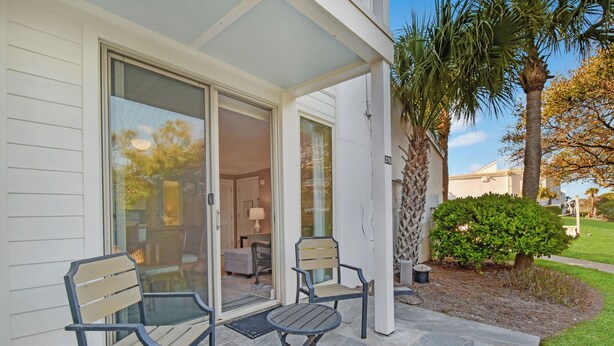 A patio with two chairs and a table, a sliding glass door leading to an interior space, and a small tree and bush in the yard. - Villa 376 - Driftwood Cottage (Jekyll Island)