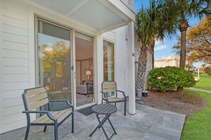 Une terrasse avec deux chaises et une table, une porte-fenêtre coulissante donnant sur l’intérieur, et un petit arbre et un arbuste dans le jardin.