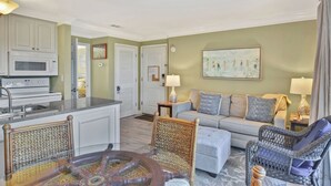 A living room with a sofa, armchairs, a coffee table, and a dining area with a kitchen counter. - Villa 376 - Driftwood Cottage (Jekyll Island)