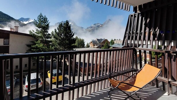 A balcony with a wooden chair, a view of a mountain, and parked cars in the background.