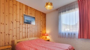A bedroom with a wooden paneled wall, a bed with a pink bedspread, a nightstand with a lamp, and a window with curtains.