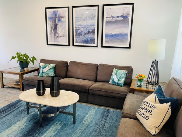 A modern living room with a brown sofa, a round coffee table, and a blue striped rug. There are three framed abstract paintings on the wall, a potted plant, and a small side table with a lamp and flowers. - Moonlight Retreat, near Wedding Venue & Breweries (Dripping Springs)