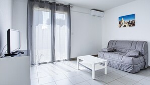 A modern living room with a grey sofa, a white coffee table, a flat-screen TV on a stand, and a large window with curtains.