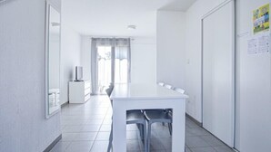 A bright, spacious room with a large white table and chairs, a television on a low cabinet, and a door with a poster.