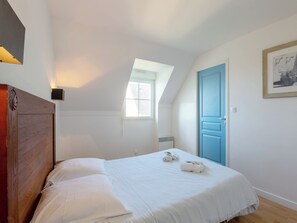 Ein Schlafzimmer mit einem Holzkopfteil, einem Bett mit weißen Leinentüchern, einer blauen Tür, einem kleinen Fenster und einer wandmontierten Leuchte.