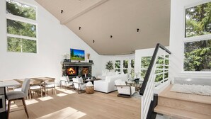 A spacious living area with a fireplace, white furniture, and large windows offering a view of greenery.