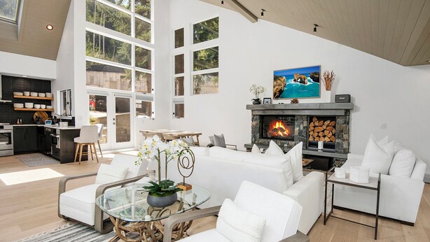 A spacious living room with a fireplace, white sofas, a glass coffee table, and a kitchen area with a large window.