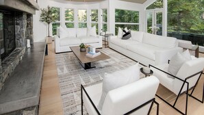 A modern living room with a fireplace, white sofas, a coffee table, and large windows offering a view of the outdoors.