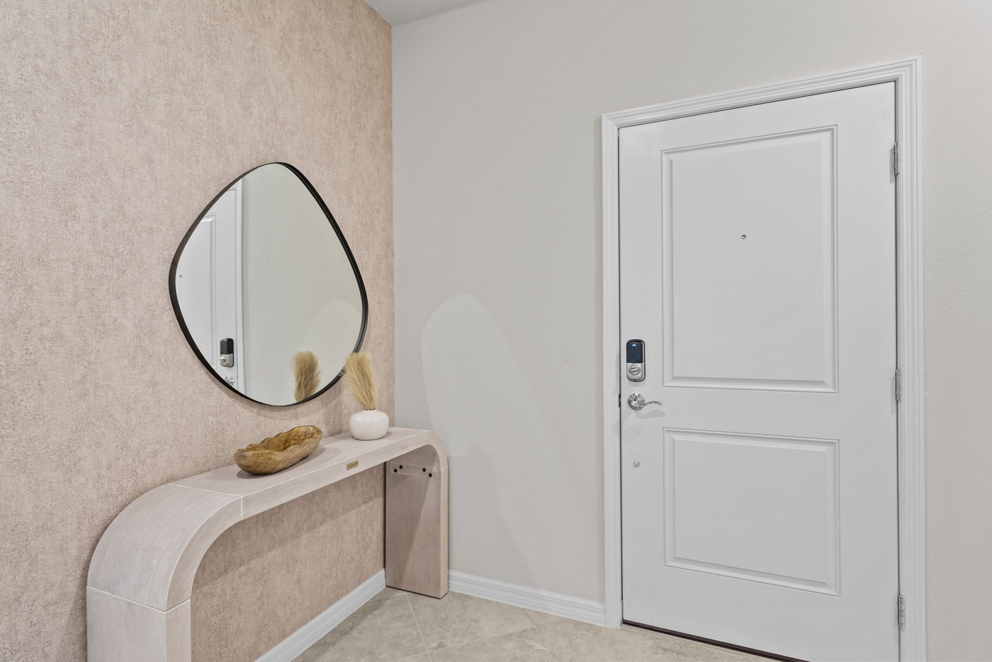 Ein Badezimmer mit einer beige strukturierten Wand, einer weißen Tür, einem Spiegel mit schwarzem Rahmen und einem Waschtisch mit heller Arbeitsplatte.