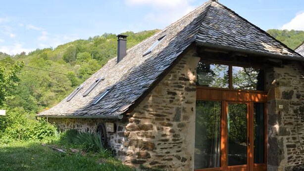 A stone cottage with a sloped roof, a chimney, and a glass door leading to a wooden interior.