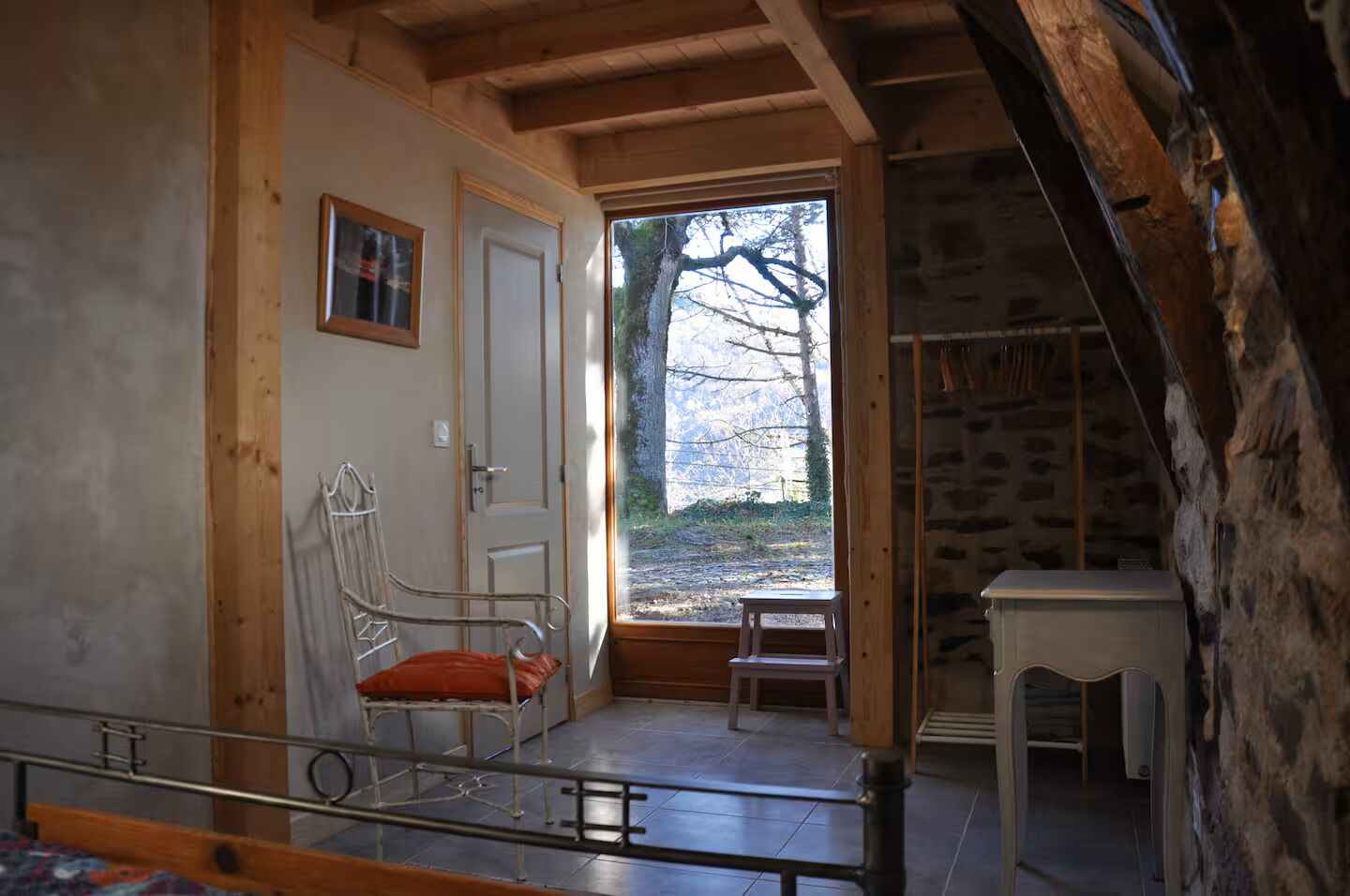 Een landelijke kamer met een houten plafond, een metalen stoel met kussen, een klein tafeltje en een deur die naar buiten leidt.