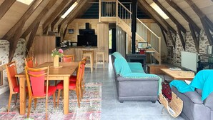 A spacious loft with a high ceiling, wooden beams, a kitchen area, a dining table with chairs, a sofa, and a staircase leading to an upper level.