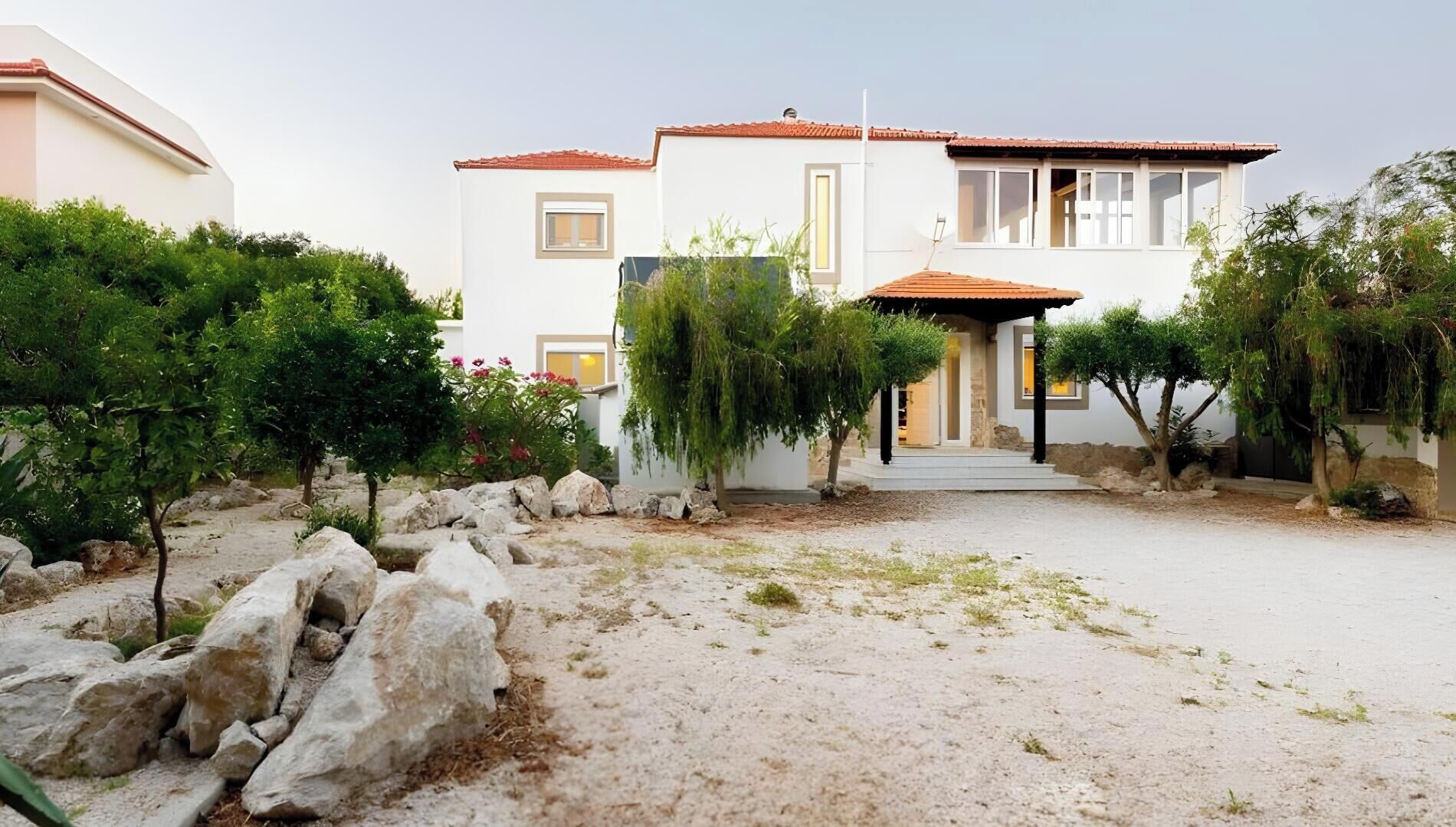 Une maison à deux étages, à l’extérieur blanc et au toit recouvert de tuiles rouges, entourée de verdure et de rochers.