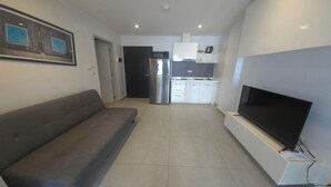 Een moderne woonkamer met een grijze bank, een flatscreen televisie bevestigd aan een witte standaard, en een keuken met witte kasten en roestvrijstalen apparatuur.