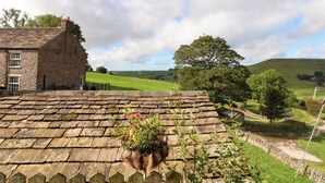 A stone cottage with a sloped roof, a hanging flower pot, and a view of rolling hills and a tree. - The Old Shippon (Macclesfield)
