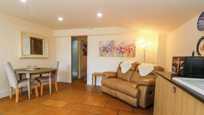 A living room with a brown leather sofa, a dining table with chairs, a kitchen area with a stove, and a painting on the wall. - The Old Shippon (Macclesfield)
