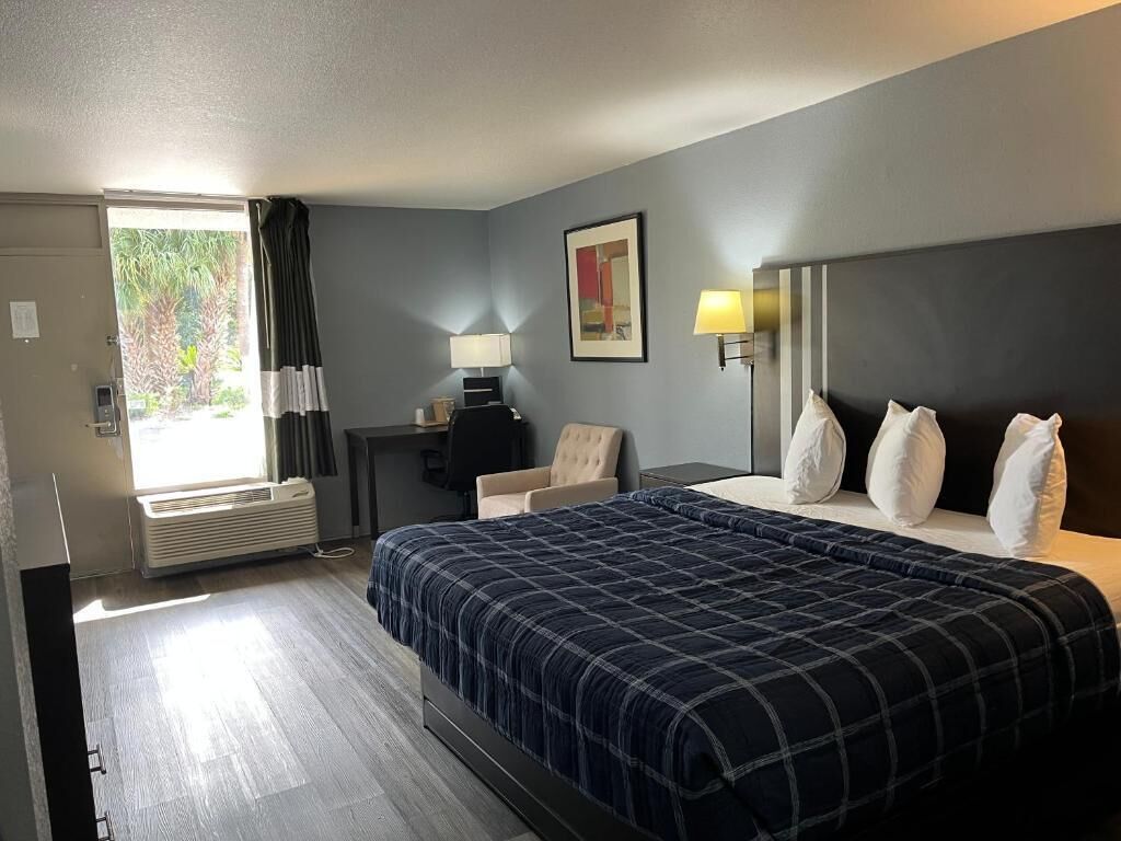 A hotel room with a bed, a desk, a chair, a lamp, and a window.