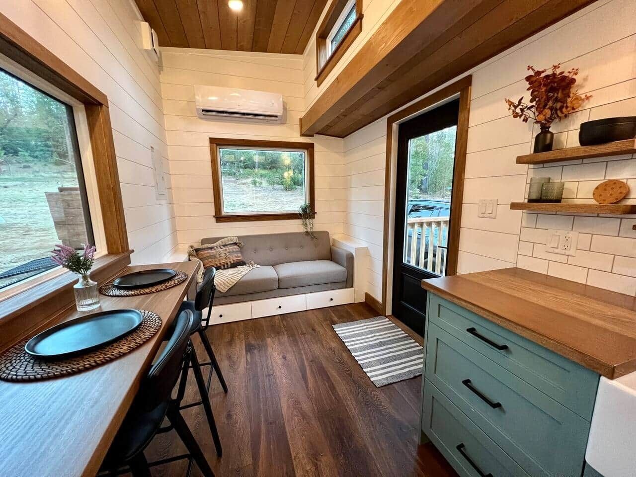 A compact kitchen with a dining area, a sofa, and a window overlooking a natural landscape.