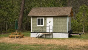 Une petite cabane avec une seule porte et une seule fenêtre, une terrasse en bois et une table de pique-nique avec un parasol.