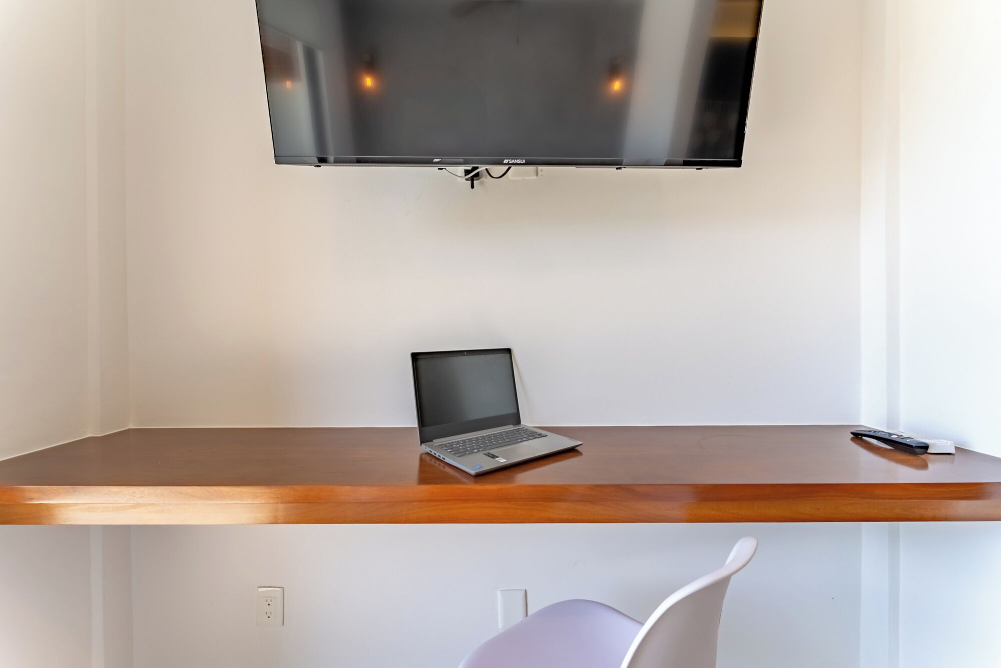 Une pièce avec un bureau en bois, un ordinateur portable, un téléviseur fixé au mur et une chaise blanche.