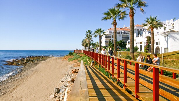 A wooden boardwalk along a beach with palm trees and white buildings in the background. - Seaview apartment in El Porton Calahonda (Sitio de Calahonda)
