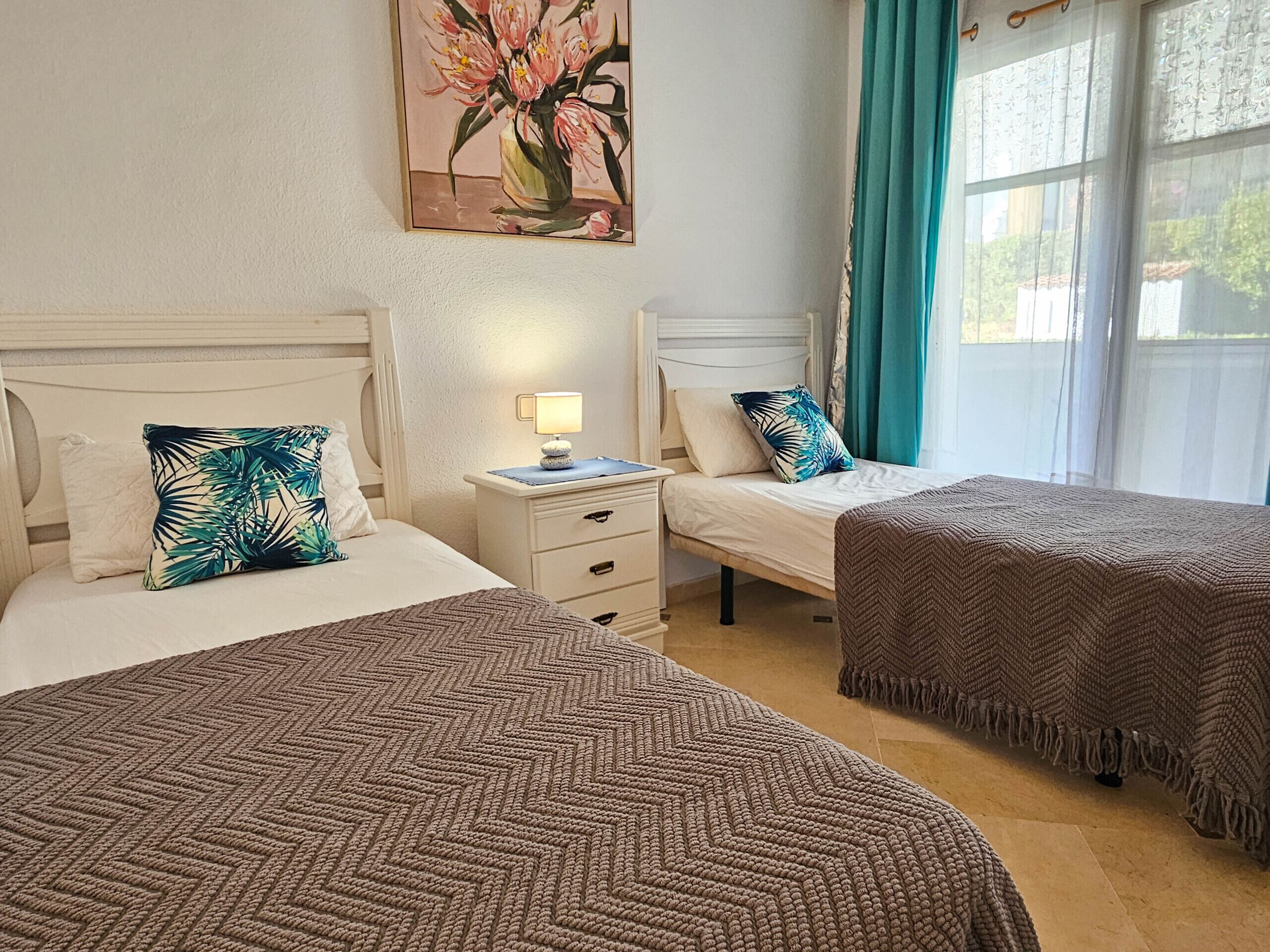 Une chambre d’hôtel avec deux lits, une table de chevet, une lampe, un tableau et une fenêtre avec des rideaux.
