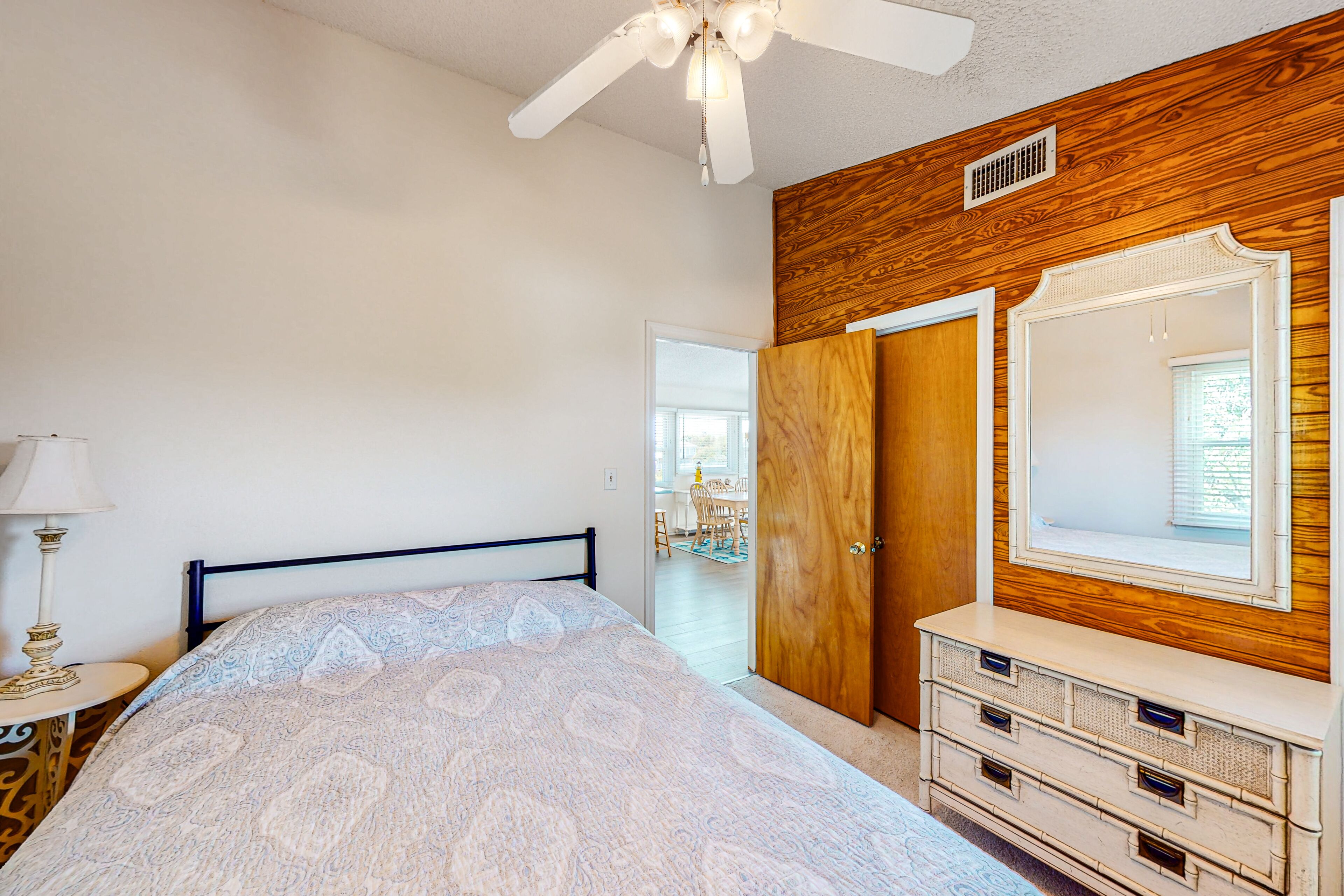 A bedroom with a bed, dresser, mirror, ceiling fan, and a door leading to another room.