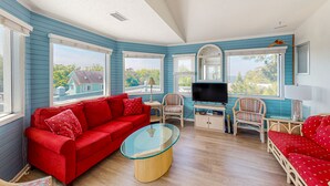 A living room with a red sofa, a glass coffee table, wicker chairs, a TV, and large windows with a view of houses. - 4BR Soundview | Deck | Washer/Dryer (Duck)
