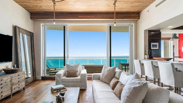 A spacious living room with a large sofa, two armchairs, and a coffee table. There is a wooden ceiling and a large window offering a view of the ocean. - Stunning Oceanfront Penthouse at the Ritz-Carlton on Singer Island (Riviera Beach)
